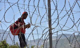 Spanish city of Ceuta in crisis with influx migrants epa09209507 A migrant waits to sneak through the fence in the northern town of Fnideq in an attempt to cross the border from Morocco to the Spanish enclave of Ceuta, in North Africa, 18 May 2021. At least 5,000 migrants, an unprecedented influx in Spanish authorities said a period of high tension between Madrid and Rabat crept into Ceuta on 17 May, a record for a day. They reached the coast by swimming or walking at low tide from beaches a few kilometers to the south, some using inflatable swimming rings. EPA/JALAL MORCHIDI